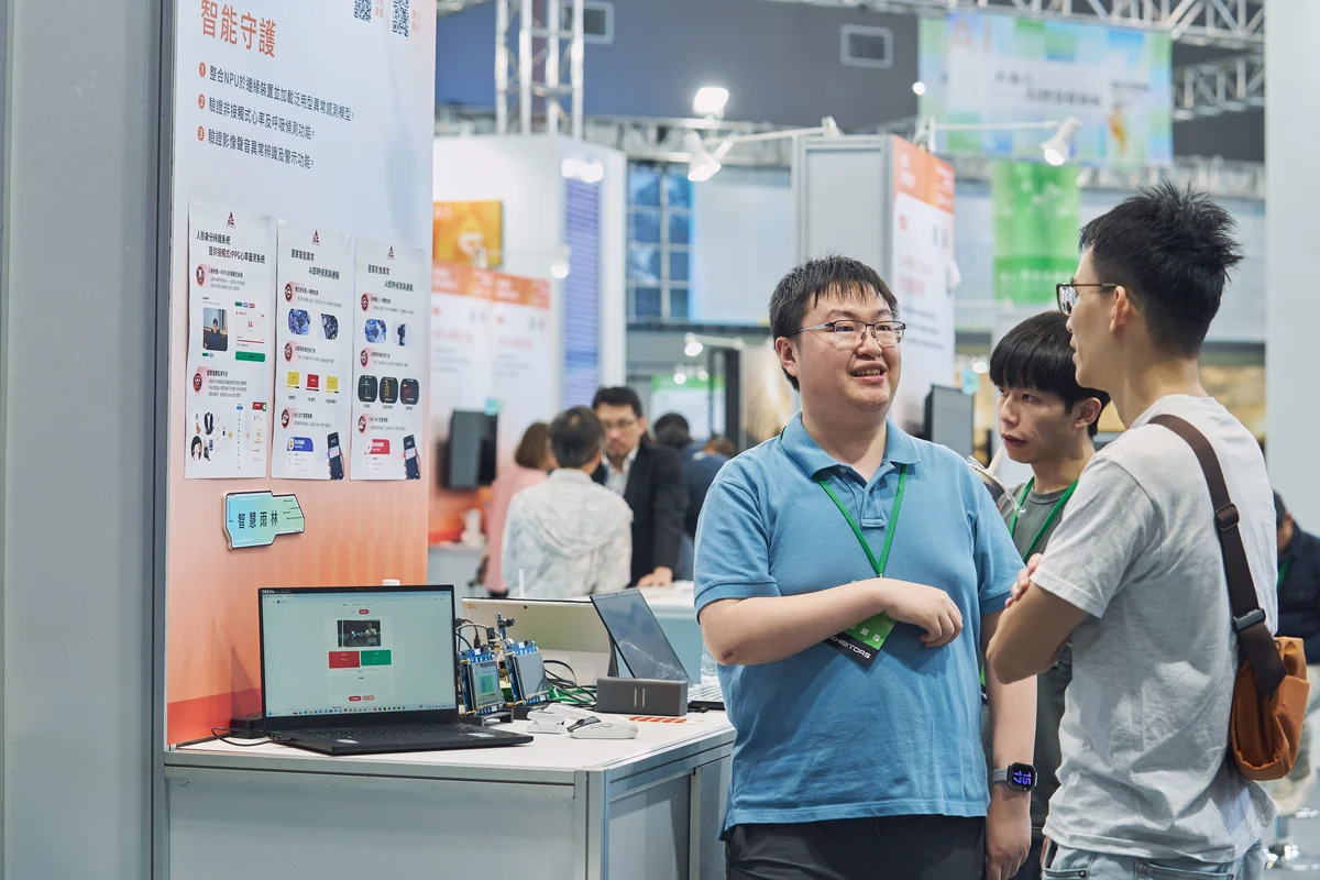 An exhibitor talks with a visitor beside a laptop and demonstration hardware at the Smart Care booth.