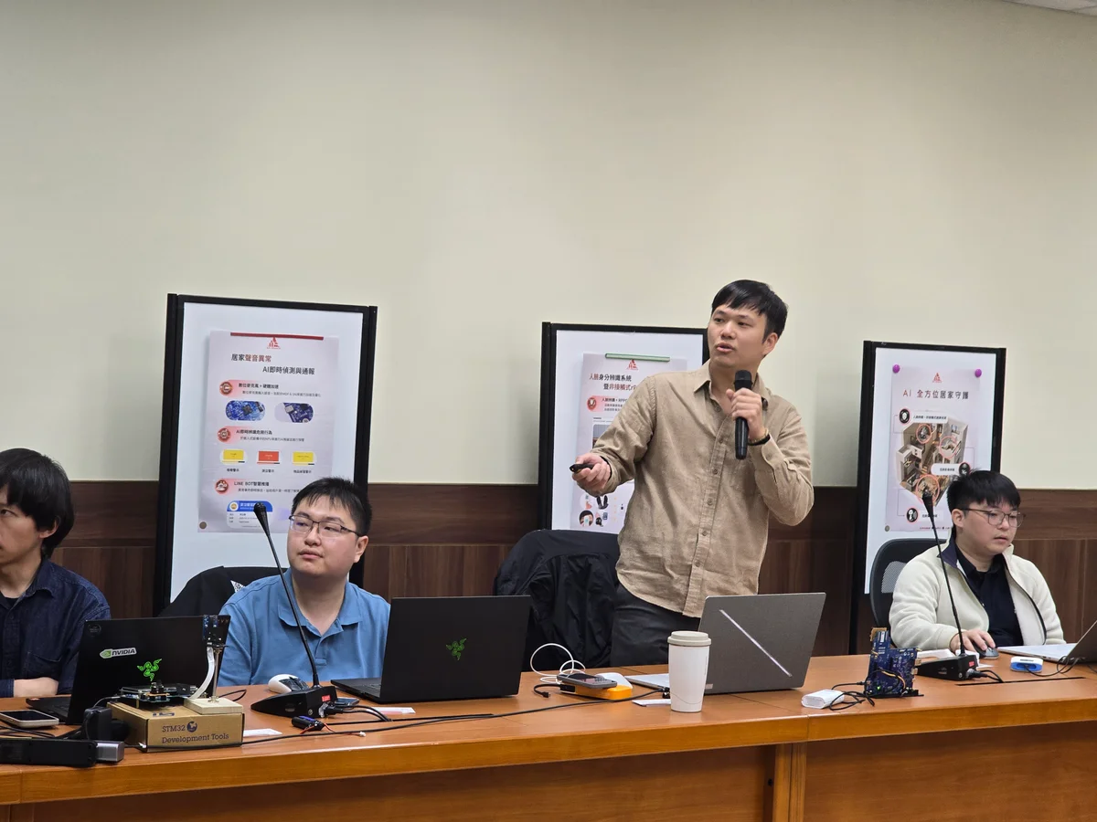 A speaker holding a microphone shares ideas beside colleagues and demonstration equipment at a meeting table.