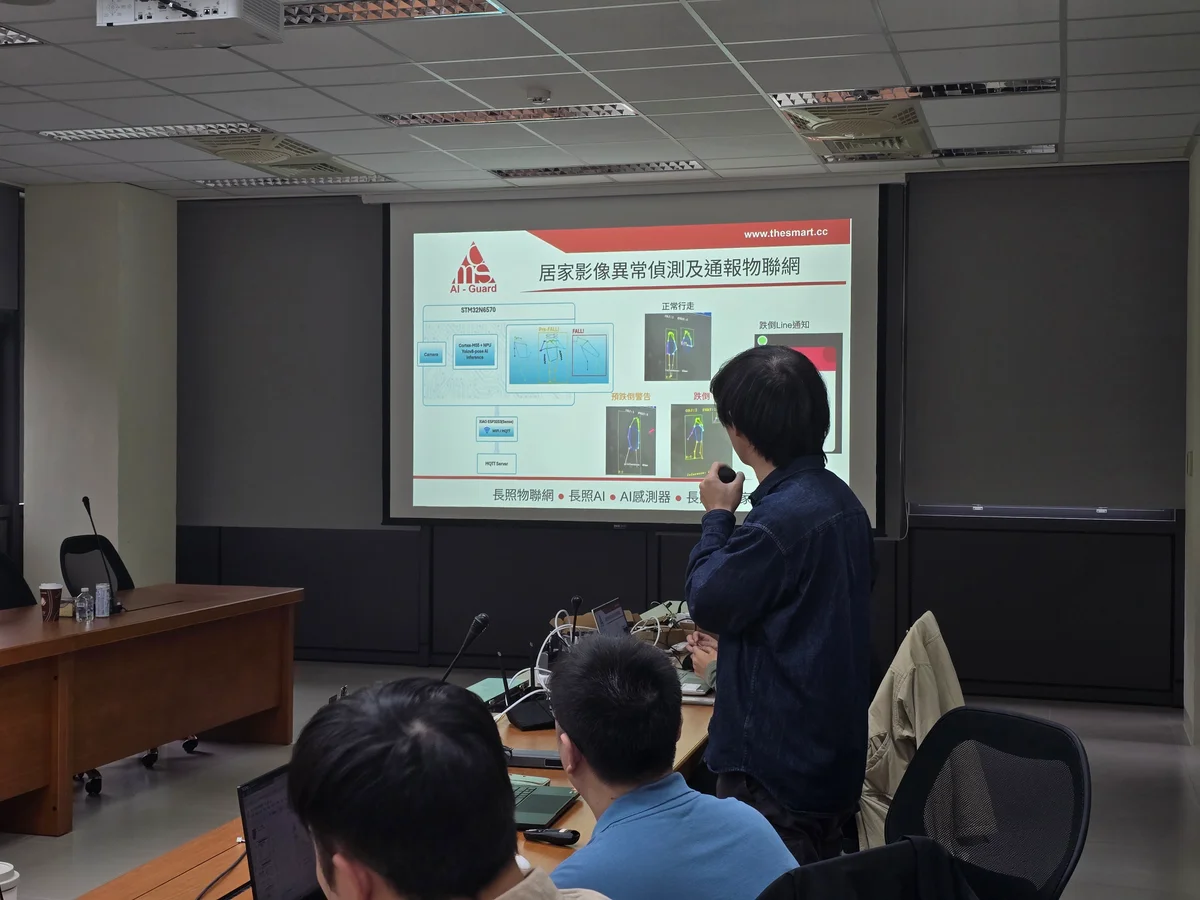 A presenter explains a projected camera detection and notification workflow to people seated in a meeting room.